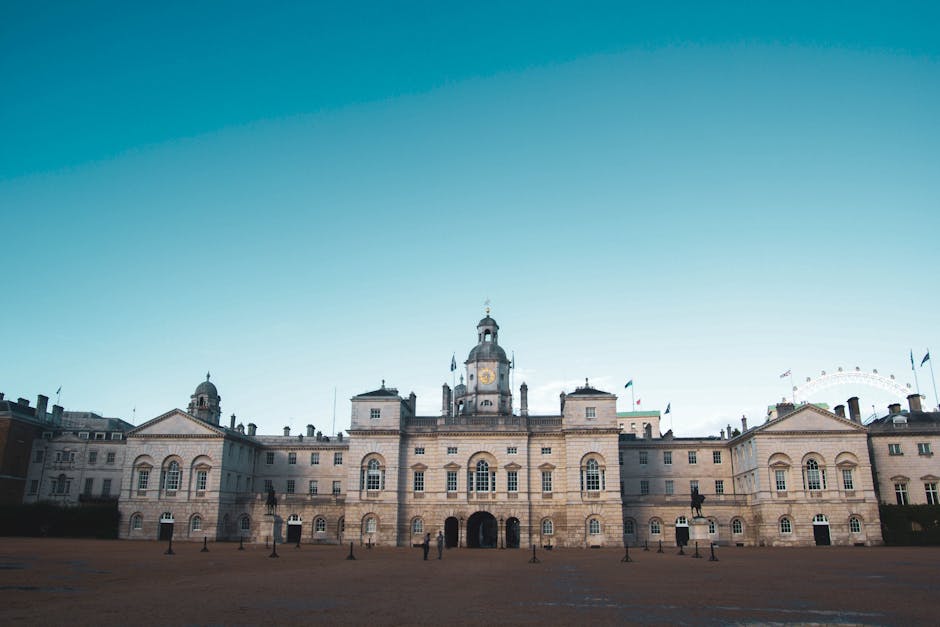 Photograph of the exterior of the Royal Hospital Chelsea, showing the historic, classical-style building with large columns, ornate architectural details, and a series of tall windows. The building is