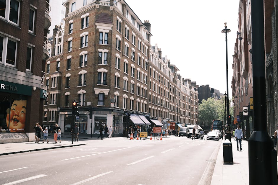 A row of elegant white multi-storey Victorian townhouses with ornate black wrought iron balconies and decorative columns, situated on a tree-lined street in Chelsea. Several cars, including a silver e